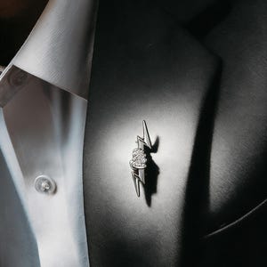 Könnte beinhalten: Ein silberfarbener Blitz-Reversnadel mit einem kleinen Blumendesign in der Mitte. Die Nadel ist an einem schwarzen Blazer über einem weißen Hemd befestigt. Die Nadel ist etwa 3,8 cm lang.
