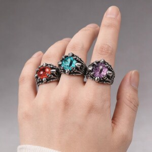 Puede incluir: Tres anillos ornamentados con grandes gemas de colores se muestran en una mano. Los anillos presentan un engaste de metal oscuro con un diseño texturizado y detalles puntiagudos. Las gemas son rojas, turquesas y moradas.