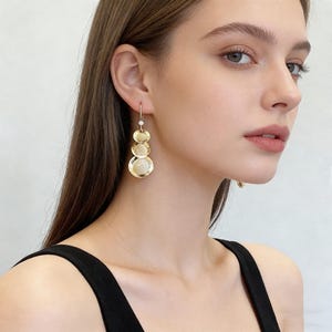 Könnte beinhalten: Goldfarbene Ohrhänger mit drei abgestuften kreisförmigen Elementen. Die Ohrringe haben ein zartes Design mit einem kleinen, runden Akzent oben. Die Ohrringe werden an einer Person gezeigt.