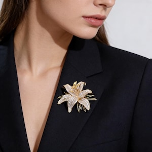 May include: A gold-toned lily flower brooch with sparkling crystals. The brooch is pinned to a black blazer. The flower has detailed petals and a delicate design, creating an elegant accessory.