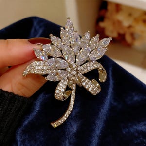 Puede incluir: Broche dorada con forma de flor estilizada. La broche presenta un grupo central de cristales transparentes en forma de lágrima, rodeados de piedras más pequeñas y brillantes. Un lazo y un tallo delicados completan el diseño floral.
