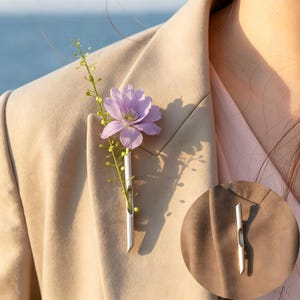 Könnte beinhalten: Ein silberner Revers-Anstecker mit schlankem, röhrenförmigem Design, der einen Zweig mit Grün und eine hellviolette Blume hält. Der Anstecker ist an einem beigen Blazer befestigt, mit einer Nahaufnahme des Ansteckers in einem kreisförmigen Einsatz.