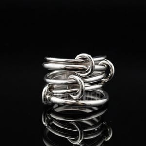Peut inclure: Une bague argentée au design unique. La bague présente plusieurs anneaux reliés par de petits maillons incurvés. Le métal poli reflète la lumière, créant un aspect brillant et accrocheur. La bague est présentée sur fond noir.