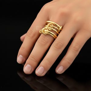 Op de afbeelding: Een goudkleurige ring met een knoopontwerp. De ring heeft drie banden, elk met een knoopachtig detail. De ring wordt getoond op een hand met een zwarte achtergrond.
