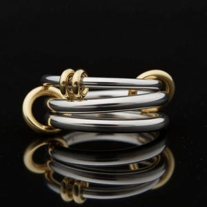 Könnte beinhalten: Ein moderner Ring mit einem Design aus gemischten Metallen. Der Ring kombiniert Silber- und Goldtöne mit mehreren ineinandergreifenden Bändern. Goldene Details sind an den Außenkanten und Verbindungselementen zu sehen. Der Ring wird auf einer reflektierenden schwarzen Oberfläche präsentiert.