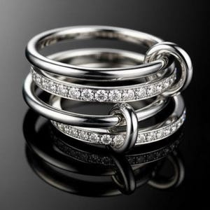 May include: Three silver rings, two with a row of small, clear gemstones, are stacked together. The rings are interlocked with a knot design. The background is black, creating a high-contrast image.