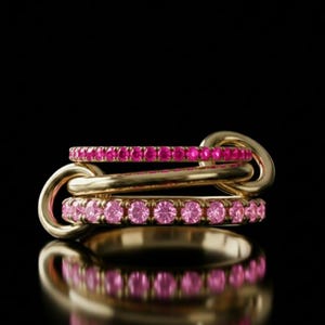 Può includere: Tre anelli d'oro collegati tra loro, ciascuno ornato da una fila di pietre preziose rosa. Gli anelli sono collegati da maglie d'oro, creando un design unico. L'immagine è ambientata su uno sfondo nero, con il riflesso degli anelli visibile sotto.