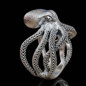 Peut inclure: Bague argentée en forme de poulpe, avec des tentacules détaillés et une tête texturée. La bague est posée sur un fond noir, mettant en valeur le design complexe et l'éclat métallique. Les tentacules de la bague s'enroulent autour de l'anneau.