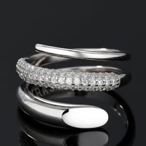 May include: A silver-toned ring with a wrap-around design. The middle band is encrusted with small, sparkling stones. The ring has a smooth, polished finish and a modern, elegant aesthetic. The ring is displayed against a dark background.