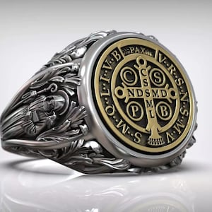 Puede incluir: Anillo plateado con centro dorado que presenta un diseño de cruz e inscripciones latinas. El anillo tiene intrincados detalles florales y religiosos, con una figura en el lateral. El anillo es de estilo sello.