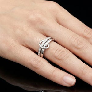 May include: A silver ring with a knot design. The ring has a band adorned with small, clear stones. The knot design is the focal point, giving the ring a unique and elegant appearance. The ring is displayed on a person's finger.