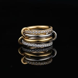 Könnte beinhalten: Ein Stapel von fünf Ringen in Gold- und Silbertönen. Zwei Ringe sind goldfarben mit einem Band aus kleinen, klaren Steinen. Zwei Ringe sind silberfarben und einer ist goldfarben. Die Ringe sind auf einer reflektierenden schwarzen Oberfläche gestapelt.