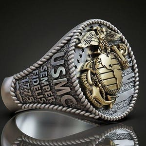 Op de afbeelding: Zilveren en gouden ring met het USMC-embleem, met een adelaar, wereldbol en anker. De ring heeft de tekst "USMC Semper Fidelis 1775" en een touwachtige rand. De ring wordt getoond op een reflecterend oppervlak.