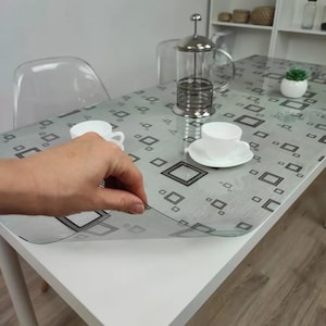 Könnte beinhalten: Ein weißer rechteckiger Tisch mit einer grau gemusterten Tischdecke. Eine Hand hebt eine Ecke der transparenten Kunststofftischdecke an. Eine French Press, weiße Tassen und eine kleine Topfpflanze stehen auf dem Tisch.