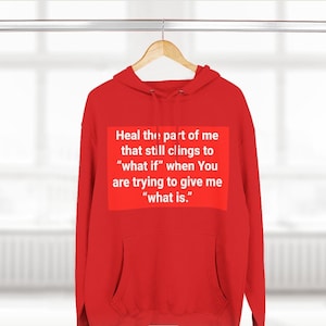 May include: A red hoodie with a white rectangular graphic containing the text: "Heal the part of me that still clings to 'what if' when You are trying to give me 'what is.'" The hoodie is hanging on a wooden hanger.