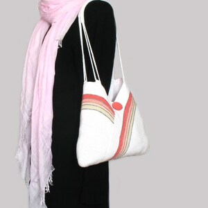 May include: A white tote bag with a rainbow stripe design and a red button. The bag has two white straps and is being held by a person wearing a black dress and a pink scarf.