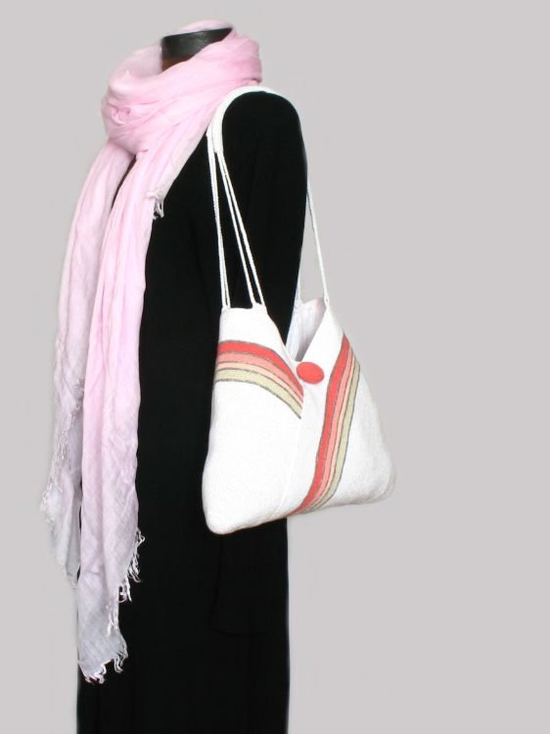 May include: A white tote bag with a rainbow stripe design and a red button. The bag has two white straps and is being held by a person wearing a black dress and a pink scarf.