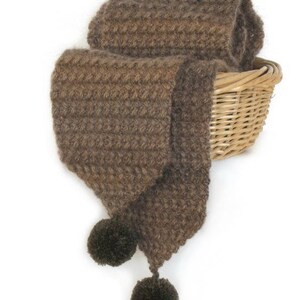 May include: A brown knitted scarf with two large pom-poms on the ends. The scarf is folded and placed in a wicker basket.