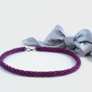 May include: A purple beaded necklace with a silver clasp, lying on a white surface with a silver satin bow in the background.