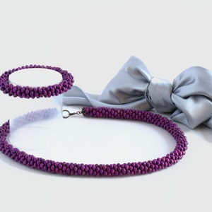 May include: A purple beaded necklace and bracelet set. The necklace is longer and has a clasp. The bracelet is shorter and does not have a clasp. The set is displayed on a white surface with a silver ribbon.