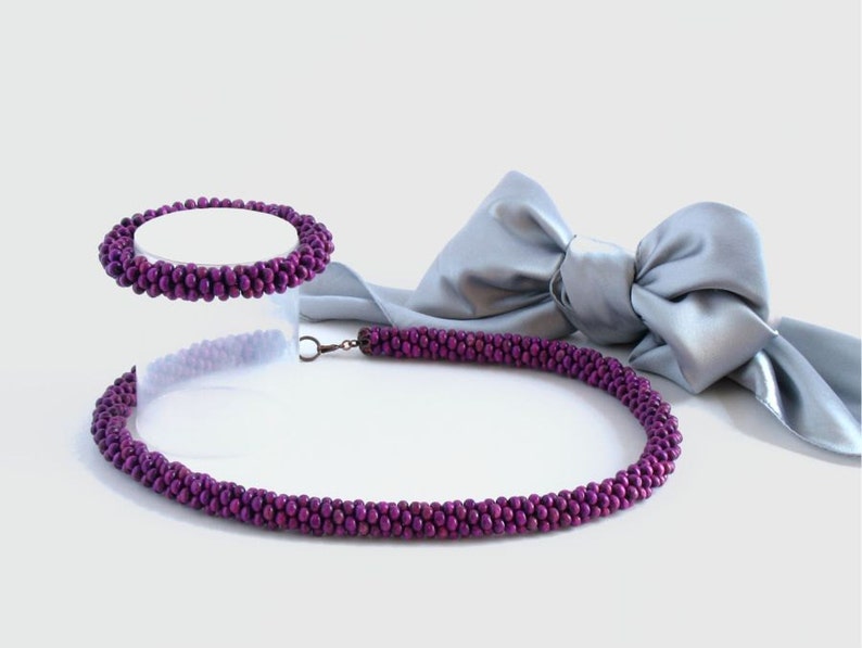 May include: A purple beaded necklace and bracelet set. The necklace is longer and has a clasp. The bracelet is shorter and does not have a clasp. The set is displayed on a white surface with a silver ribbon.
