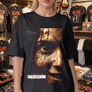 May include: A black short-sleeve t-shirt featuring a large, detailed graphic of a human face with a textured, weathered appearance. The word OBSESSION is printed in bold white letters below the face, with smaller text above the graphic.