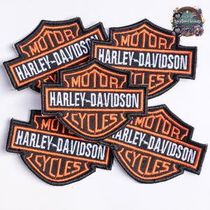 Peut inclure: Un ensemble de cinq écussons brodés arborant le logo Harley-Davidson. Chaque écusson en forme de bouclier présente un fond noir, des bordures orange et le texte blanc MOTOR, HARLEY-DAVIDSON et CYCLES disposé en couches superposées.