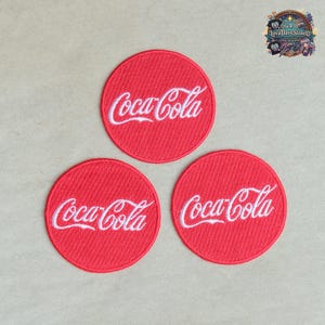 Może przedstawiać: Trzy okrągłe haftowane naszywki, każda z żywym czerwonym tłem i białym logo Coca-Cola wyhaftowanym na środku. Naszywki mają wyraźną diagonalną fakturę materiału i czyste, zaokrąglone krawędzie, prezentujące kultowy napis.