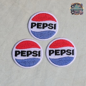 Può includere: Tre toppe ricamate circolari con il logo Pepsi. Ogni toppa presenta una sezione superiore rossa, una fascia centrale bianca con il testo nero PEPSI e una sezione inferiore blu, il tutto racchiuso da un bordo bianco cucito su tessuto.