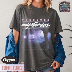 Op de afbeelding: Een antracietgrijs t-shirt met korte mouwen en de tekst UNSOLVED mysteries in wit en lichtblauw. De afbeelding toont een donker, mistig bos met een Dead End-bord. Onder de afbeelding staat de tekst GHOSTS HAUNTINGS THE UNEXPLAINED.