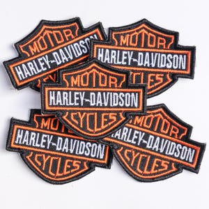 May include: Five embroidered patches featuring the Harley-Davidson Motor Cycles logo. Each patch is shield-shaped with a black background, orange border, and white text that reads MOTOR, HARLEY-DAVIDSON, and CYCLES in a stacked, layered arrangement.