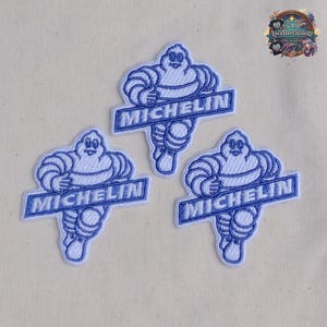 Puede incluir: Tres parches bordados que muestran al personaje Michelin en blanco con contornos azules. Cada parche representa la figura con los brazos cruzados, situada sobre una banda rectangular azul que contiene el texto blanco MICHELIN.