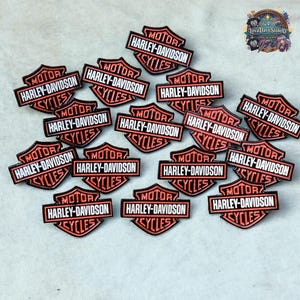 May include: A collection of shield-shaped Harley-Davidson pins featuring a black and orange colour scheme. Each pin displays the text Harley-Davidson across the centre bar with the word Motor above and Cycles below, arranged in a scattered pattern.