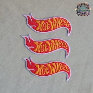 Puede incluir: Tres parches bordados con el logotipo de Hot Wheels en forma de pancarta ondulada. Cada parche tiene un fondo rojo brillante con letras amarillas estilizadas que dicen Hot Wheels, todo delineado con un borde blanco fino para contraste.