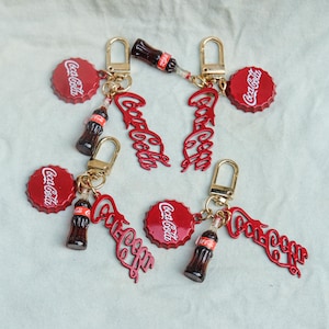 Pode incluir: Quatro porta-chaves com o tema Coca-Cola, cada um com um fecho lagosta dourado, um pendente de caricas vermelha com texto branco, uma miniatura de garrafa de vidro castanho escuro e um recorte de metal vermelho do logótipo Coca-Cola.