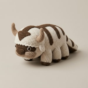 May include: A handmade crochet plush toy of a bison-like creature. The toy features a cream-colored body with brown markings on the head and back. It has small, curved horns and dark eyes. The toy is approximately 10 inches long.