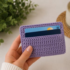 May include: A rectangular lavender crochet card holder featuring a textured stitch pattern. The holder contains two stacked credit cards, with the top card displaying a blue background, a yellow circular icon, and a contactless payment symbol.