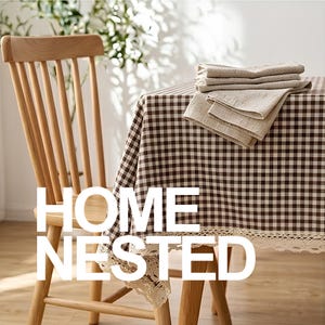 May include: A wooden chair with vertical slats stands beside a table covered in a brown and white checkered tablecloth featuring a delicate cream-coloured lace trim along the edge. A stack of folded beige napkins rests on the table surface.