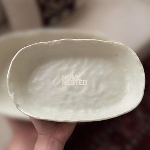 May include: An oval-shaped off-white ceramic dish with an irregular, hand-formed rim and a textured, wavy interior surface. The center of the dish features the white text HOME NESTED printed in a clean, sans-serif font against the glossy finish.