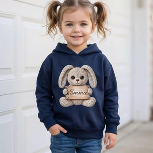 May include: A navy blue hooded sweatshirt featuring a knitted bunny holding a wooden sign with the name "Emma". The bunny has large ears and a sweet expression. The child is wearing blue jeans.