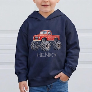 May include: Navy blue hooded sweatshirt featuring a red monster truck graphic. The truck has large tires and is labeled "HENRY" in gray text. The sweatshirt is made of a soft material and has a hood and a front pocket.