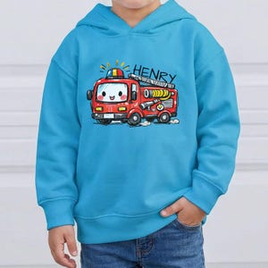 May include: A turquoise blue hoodie with a cartoon fire engine design. The red fire engine has a smiling face and the name "HENRY" above it. The sweatshirt is made of a soft material and has a kangaroo pocket.