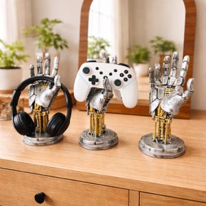 Puede incluir: Tres soportes con forma de mano robótica. Uno sostiene auriculares negros, otro un mando de videojuegos blanco y el tercero está vacío. Los soportes tienen manos plateadas, mecanismos internos dorados y bases redondas grises. Los objetos están sobre una superficie de madera.