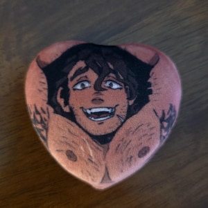 Könnte beinhalten: Herzförmiges Plüschtier mit einer Cartoon-Illustration eines lächelnden Mannes mit dunklem Haar und muskulöser Brust. Das Plüschtier hat einen pfirsichfarbenen Hintergrund und das Gesicht des Mannes ist detailliert mit offenen Augen und einem breiten Grinsen. Tätowierungen sind auf den Armen sichtbar.