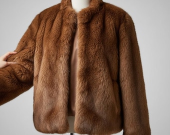 May include: A brown faux fur jacket with a round collar. The jacket is displayed on a mannequin. The jacket has a soft, textured appearance and a warm, inviting color. The jacket is open in the front.