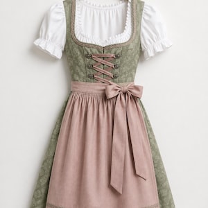 May include: A dirndl dress featuring a sage green bodice with a subtle floral pattern, a white blouse with short puffed sleeves, and a solid dusty pink apron. The bodice includes decorative metal hooks with a pink ribbon lace-up front closure.