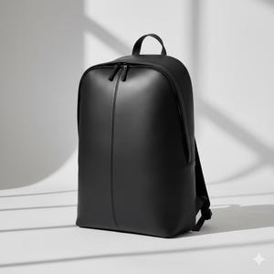Könnte beinhalten: Ein schwarzer Rucksack mit abgerundeter Oberseite und kleinem Griff. Der Rucksack hat eine vertikale Naht auf der Vorderseite und zwei Reißverschlüsse. Die Tasche besteht aus einem glatten Material und steht auf einer weißen Oberfläche.