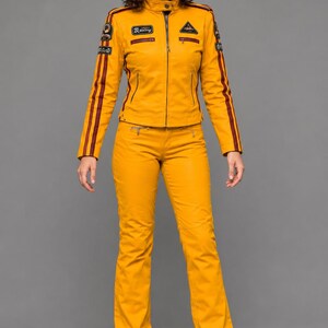 May include: A mustard yellow jacket and matching pants set. The jacket features a stand-up collar, a front zipper, and dark red stripes along the sleeves. Multiple embroidered patches are visible on the chest and sleeves. The pants are straight-leg.