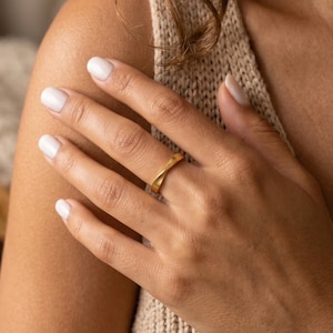 May include: A gold ring with a distinctive crossed design. The ring is on a person's finger, captured in a close-up shot. The ring's design is simple but elegant, and it appears to be made of a shiny metal.