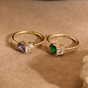Op de afbeelding: Twee gouden ringen, elk met een hartvormige edelsteen. Eén ring heeft een paarse en heldere steen, de andere een groene en heldere steen. De ringen worden gepresenteerd op een neutraal getinte ondergrond.